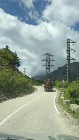 A truck carrying a large piece of industrial equipment, possibly a generator or transformer, drives along a winding mountain road. The road is paved and has a white line dividing the lanes. To the left of the road, there is a steep, green, tree-covered mountainside. To the right, there are more power lines supported by tall metal towers, and a signpost with an arrow indicating a turn. In the background, more mountains can be seen, some shrouded in clouds. The sky is a vibrant blue with large, puffy white clouds, suggesting it is daytime and the weather is fair, though perhaps with a chance of rain given the clouds. The overall scene depicts a journey through a scenic, mountainous region, likely in Russia given the provided location context. The industrial equipment being transported suggests construction or maintenance work on the power grid in this remote area. There is no discernible text on the objects or structures, apart from the signage which is not legible. The atmosphere is one of travel and progress through a rugged natural landscape.