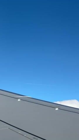 The video shows a view from an airplane window, focusing on the wing of the plane and the blue sky. A white contrail from another aircraft is visible in the distance. The tail of the plane is blue with a white logo on it. The weather appears clear.