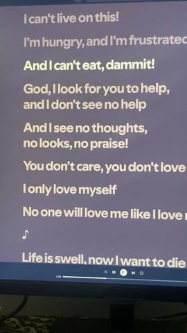The video shows a close-up of a computer monitor displaying the lyrics of a song on what appears to be a music streaming platform. The lyrics are written in white text on a purple background. The song appears to be titled "God" as it's a recurring word in the lyrics, and it includes themes of frustration, loneliness, and despair. The playback controls of the music player are visible at the bottom of the screen, along with a progress bar. The screen is being viewed from a slightly angled perspective. There is a bed with a patterned bedspread visible below the monitor.