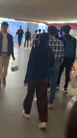 The video shows a crowded indoor public space, possibly a subway station or underground pedestrian passage. People of various ages and ethnicities are seen walking in different directions, creating a dynamic, blurred scene. Individuals' clothing styles vary, including casual attire like jackets, jeans, and sneakers. Some people carry bags or backpacks, suggesting travel or shopping. Brightly colored windows or wall panels are visible in the background, adding visual interest. The floor is light grey or beige, and the lighting appears artificial. The camera moves along with the crowd, adding to the sense of constant motion.