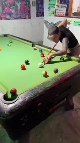 A woman plays pool in a dimly lit indoor room with graffiti-like artwork on the walls. She takes a shot, then turns quickly and walks away out of frame. The camera follows her to an outdoor setting, where she stands by a wooden object, possibly a boat, laughing. The lighting is low, indicating it is nighttime. She is wearing a bikini top and a sarong.