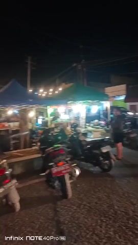 The video shows an outdoor night market scene. A line of parked motorcycles is visible in the foreground. In the background, there are stalls or tents illuminated by strings of warm-toned lights, creating a festive atmosphere. People are seen milling about the market, and there are tables and chairs set up, suggesting dining or seating areas. The overall impression is one of a lively and busy evening market.