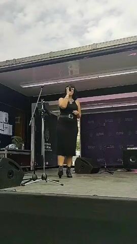 A woman sings into a microphone on stage. She wears a black, form-fitting dress and cowgirl boots. A black belt cinches her waist, and she wears a necklace. She stands on a stage with various pieces of musical equipment and a backdrop with the words "Secretaria de Cultura". The sky is overcast.