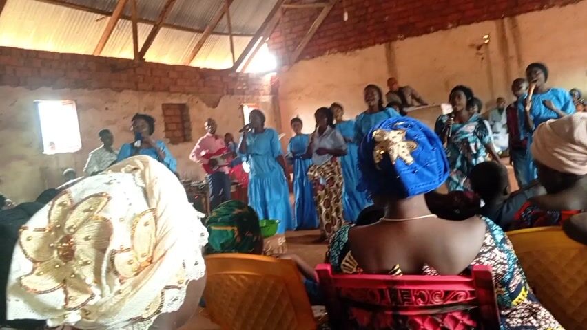 The video captures a vibrant gospel performance within what appears to be a church hall. A choir, largely composed of women dressed in light blue gowns, is actively singing and dancing on a makeshift stage. Several members hold microphones, suggesting they are leading the singing.  A guitarist in a pink shirt and other musicians are to the side, providing accompaniment. In the foreground, viewers see the backs of seated audience members, some wearing colorful headwraps.  The hall itself is simple, with brick walls, a wooden roof, and limited natural light filtering through small windows.  A man in a suit stands on a platform in the background, observing the performance.  Overall, the scene conveys a sense of lively religious expression and community gathering.