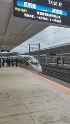 The video shows a high-speed train at a station platform. The train is white with red and gray stripes, and passengers are standing near the doors with luggage. A digital display above shows train information, including the train number G1380, the destination 昆明南 (Kunming South), and the departure time 17:50. The platform is concrete and has a yellow safety line along the edge. Overhead power lines are visible. The sky is overcast.
