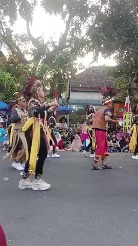 A group of people in traditional costumes are performing a dance in a street. The dancers are wearing elaborate headdresses with feathers, black tops with yellow sashes, and black leggings. Some of them are carrying props. There is a crowd of spectators on the side of the street, some of whom are seated and others are standing. In the background, there are buildings and trees. The sky is visible and appears to be overcast. One of the dancers is wearing white sneakers, and another is wearing white tennis shoes. The dancers' movements are energetic and coordinated. Some of the spectators are watching the performance attentively. The overall scene suggests a cultural festival or celebration.