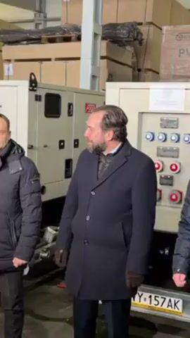 Two men stand side-by-side in an indoor space. The man on the left wears a black puffer jacket and the man on the right wears a dark overcoat and leather gloves. Both men are turned slightly to their right. Behind them are two large generators, and stacks of wooden sheets wrapped in plastic.  The generator on the right has a control panel with various switches and outlets. A license plate is visible on the bottom of the generator, with "WY" followed by numbers and letters. A microphone is visible in the bottom left corner of the frame. The lighting is bright, suggesting a warehouse or industrial setting.