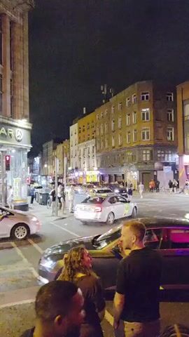 The video shows a street scene at night. Several people are standing on the sidewalk and in the street. There are cars, including taxis, on the road. Buildings line the street, some of which appear to be shops or businesses. The lighting suggests it is nighttime, with streetlights and lights from the buildings illuminating the scene. There are traffic lights visible. The atmosphere appears to be social, with groups of people conversing.