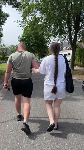 A man and a woman walk along an asphalt road. The man is muscular and wearing a green t-shirt and black shorts and shoes. The woman has long brown hair in a thick braid, and is wearing a white t-shirt, white shorts, and black sandals. She has a large black tote bag on her left shoulder. The man and woman are holding hands. They are walking away from the camera. Trees and houses are visible on either side of the road. A white RV is visible in the background. The sky is overcast.