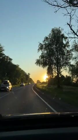 A car is traveling along a road at sunset. The sun is low in the sky, causing glare on the windshield. Trees line either side of the road. Another car is visible in the distance traveling in the same direction. The windshield has some water spots. The sky is a clear blue.