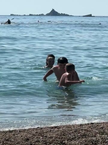 The video shows a group of children playing in the water at a beach. In the foreground, the water is clear and shows pebbles on the seabed. In the middle ground, a group of children are in the water. The two youngest children appear to be being held by an older child. One child is wearing a dark shirt and the other is wearing a darker pair of swim trunks. A girl in the background wearing a colorful swim suit is splashing water. The oldest child is on his stomach in the water, with his legs in the air, seemingly diving or jumping. In the background, there are a few people on paddleboards and some rocky islands offshore under a clear sky.