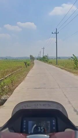 The video shows a first-person perspective of riding on a paved road. The road is lined with green fields on both sides. There are utility poles with wires running along the road. The sky is blue with scattered clouds. A digital display in the foreground shows a speed of 35, which then changes to 36. The overall environment appears to be rural or semi-rural.