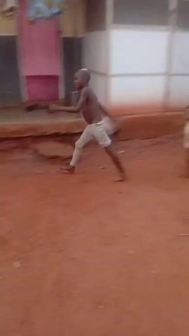 The video shows a young, shirtless child running through an outdoor, dirt-covered area. The child wears light-colored shorts and is barefoot. The child is running in a blurred manner at the beginning of the video, then the camera focuses and the child is seen running through a residential area with pink buildings and other objects. Another person in a blue shirt can be seen in the background. The ground is reddish-brown and there is green foliage present. The camera appears to be handheld and moves along with the running child. The overall setting looks like a village or rural area.