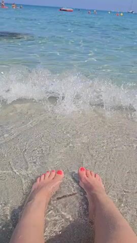 The video shows a first-person perspective of someone's feet in shallow, clear ocean water on a sunny day. The water is crystal clear, revealing the sandy bottom and small fish. Gentle waves are lapping at the person's feet, which are painted with bright pink nail polish. In the background, the blue ocean stretches to the horizon, with a few small boats and buoys visible. The overall scene is peaceful and relaxing, evoking a sense of a tropical vacation.