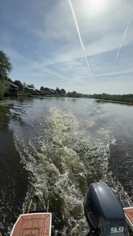 The video depicts a boat moving through a river or canal, leaving a foamy wake behind it. The boat is relatively small with a wooden deck and an outboard motor. The water reflects sunlight, causing a sparkling effect. On either side of the waterway are houses, many with well-kept gardens and docks. The sky is light blue with a few wispy clouds and what appear to be contrails. The overall atmosphere suggests a calm, sunny day.