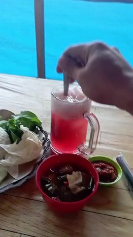 A close-up, first-person perspective video shows a hand holding a spoon and stirring a tall glass of pink liquid with ice and foam. The glass has a handle and is placed on a wooden table. To the left of the glass is a plate with white rice, green leafy vegetables, and what appears to be fried food, possibly crackers or chips. In front of the glass is a small red bowl filled with a dark stew containing meat and vegetables. To the right of the red bowl is a small green dish with red chili sauce. In the background, a blue tarp or curtain is visible. The video is short, showing only the action of stirring the drink.