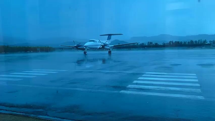 A two-propeller plane sits on a rainy airfield at dusk, viewed through a window with a blue tint. The plane faces right, with its left propeller slightly visible. White crosswalk lines are painted on the wet tarmac in the foreground. Mountains are visible in the distance, and the entire scene has a blue hue.