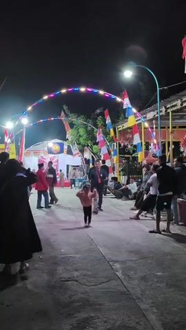 The video shows an outdoor nighttime scene with several people. In the foreground, a young child is running, followed by an adult. To the left, a person in a dark coat stands. In the middle ground, several people are gathered, some standing and some sitting. Flags are visible throughout the scene, including Indonesian flags. There are also lights strung across the area, creating an archway. In the background, there are tents and buildings. The overall atmosphere appears to be a festive or community event.