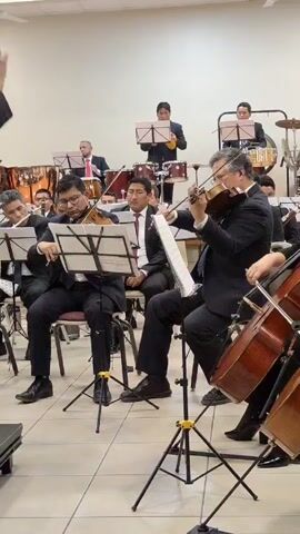 An orchestra of formally-dressed musicians is playing in a well-lit room with white walls and tiled floors. Several violin and cello players are seated in the foreground, reading sheet music on stands. Behind them, percussionists are playing drums and cymbals. Everyone appears focused and immersed in their performance.