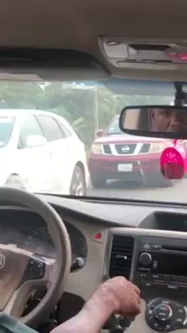 The video shows a view from inside a car, likely a Toyota minivan, driving on a paved road. The driver is visible in the rearview mirror and his hands are manipulating the gearshift and radio controls. The dashboard is visible with a Toyota logo on the steering wheel. Several vehicles are seen through the windshield, including a red Nissan Pathfinder, a white minibus with the license plate "PP478," and another red car. The environment appears to be a semi-rural area with green vegetation lining the sides of the road. A power pole is also visible on the side of the road. The overall impression is of a car driving in light traffic on a road in a developing area.