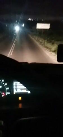 The video shows a car driving on a dark road at night. The car is moving at a speed of about 60 kilometers per hour, and there is another car in front of it. There is music playing in the car. The dashboard of the car is visible, and it is lit up. The road is asphalt, and there are some green areas on the side of the road. The sky is dark, and there are some distant lights visible.