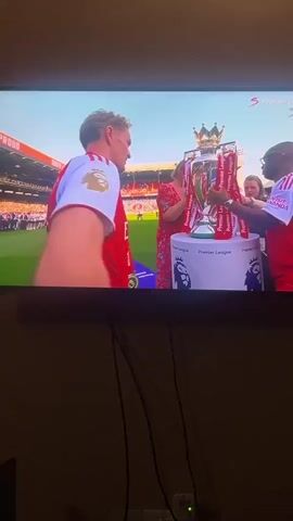 The video shows a man in a red and white jersey with a Premier League patch on the sleeve standing to the left of a Premier League trophy. Two people are holding the trophy, one on each side. The man in the jersey is looking at the trophy. The trophy is on a white pedestal with Premier League logos on it. In the background, there are other people and green grass, likely a soccer field. The scene is being displayed on a television screen. Wires dangle below the television.
