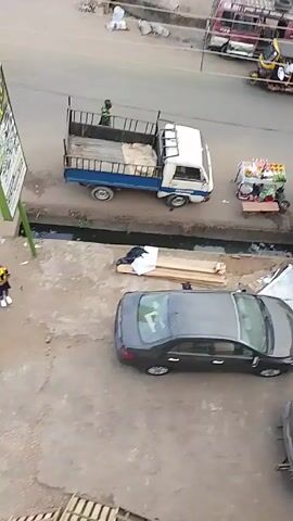 The video shows an elevated view of a street scene. A variety of vehicles are visible, including cars, trucks, and vans. A gray sedan is parked in a lot near a pile of wood. A vendor stands at a fruit cart on the sidewalk. Other structures include a metal roof and a covered vehicle. People are walking along the sidewalk.