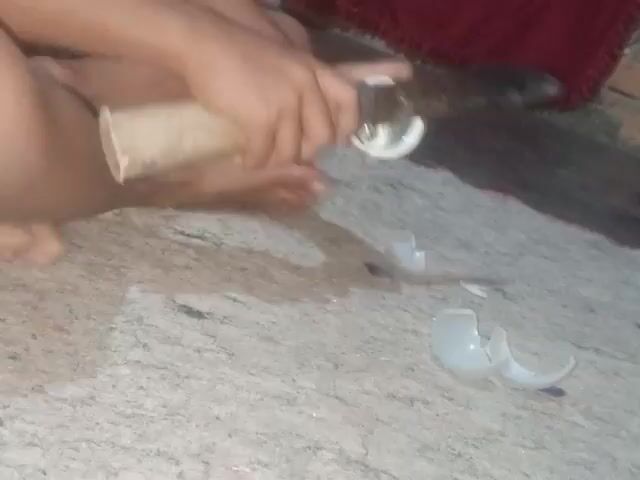 A person's hands are shown holding a large knife and a broken white object. The object appears to be a ceramic cup or bowl, with several shards scattered on a textured, light-colored surface. The person is cutting into the remaining part of the object with the knife. The person is wearing a dark t-shirt with a graphic design. A red textured mat is visible in the lower left corner of the frame. The scene appears to be indoors.