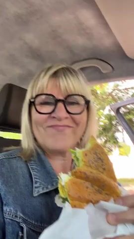 A woman with blonde hair and glasses is in a car, holding a sandwich and eating it. The car interior is visible, with a gray headliner and a door handle. Outside the car, trees and a white car are visible. The woman is looking at the camera and eating the sandwich.