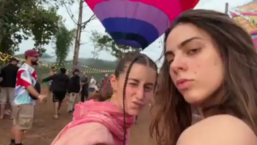 The video begins with a close-up of two young women. The woman in the foreground has long brown hair and glitter on her face. The woman behind her has her hair in pigtails and is wearing a pink scarf. The camera quickly pans to show a crowd of people standing in a field. There is a colorful hot air balloon being prepared in the background. The environment appears to be outdoor with a lot of trees. The people are mostly casually dressed. The video then ends by focusing on the colorful hot air balloon.