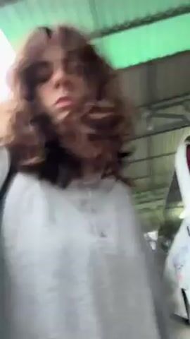 A person with long, curly brown hair is walking on what appears to be a dirt or gravel path. The person is wearing a light gray long-sleeved button-up shirt, black shorts, and black shoes. They also have a black bag slung over their shoulder. The camera angle suggests the video is being taken by the person as they walk, looking down at the ground part of the time. In the background at the start of the video, a white vehicle with red and black detailing is partially visible. The ground is covered in small rocks and some organic matter like leaves. The lighting seems somewhat dim.