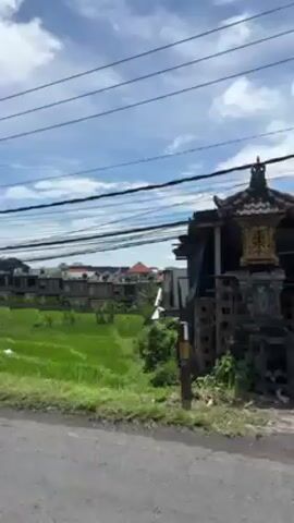 The video shows a view of a road with green fields and buildings in the background under a partly cloudy sky. Overhead power lines are visible. A person wearing a helmet and riding a white scooter drives past the camera.