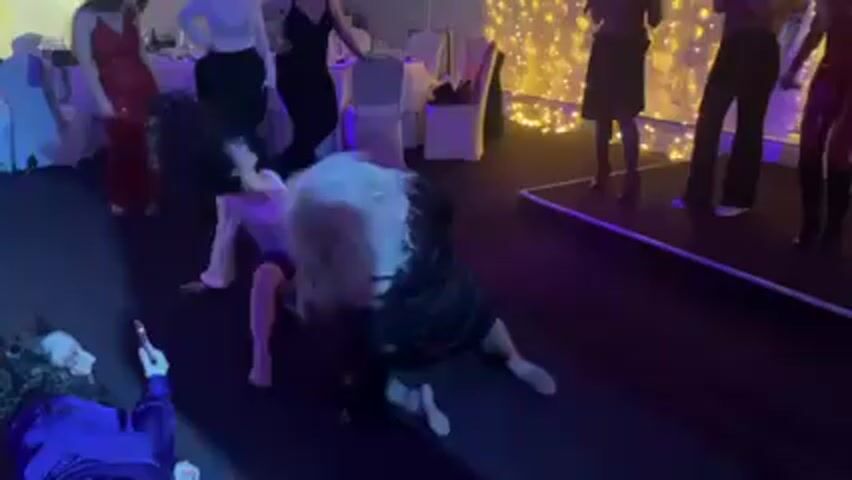 The video shows a group of women on the floor at a wedding reception, with others standing around them. There are at least three women on the floor, and they appear to be dancing and having fun. The floor is dark, and there are tables and chairs in the background. Some people are standing near a small platform. The lighting is dim, with a blue hue, and there are strings of decorative lights in the background. Several women in the background are dressed in formal attire, and one is holding a phone, likely filming. Overall, the scene appears to be a lively and informal moment at a wedding celebration.
