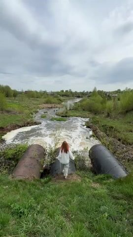 A young woman with long red hair, wearing a white top and light-colored pants, stands on a large, dark, cylindrical pipe over a flowing river. She faces away from the camera, towards the river, with her arms slightly outstretched. The river flows rapidly, creating white foam as it moves. Two other large pipes flank the central one on either side. Green grass and vegetation cover the banks of the river. In the distance, a small village is visible under a cloudy sky.