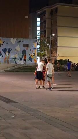 A group of young people are playing soccer in an urban square at night. The square is paved with stone tiles and lit by streetlights. In the background, there are apartment buildings and a mural on a wall. One of the players is carrying a Spanish flag draped over his shoulders. Other people are visible in the background, some sitting and some walking. The atmosphere appears to be festive and casual.
