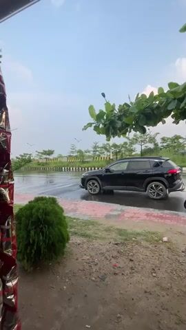 The video captures an outdoor roadside scene, likely in Purbachal, Bangladesh. A black SUV, appearing to be a Toyota Corolla Cross, is prominently parked on the side of a wet asphalt road. The ground is visibly damp, indicating recent rainfall. In the background, an auto-rickshaw, commonly referred to as a CNG in Bangladesh, drives past from left to right. Green bushes are present in the immediate foreground and along the roadside, with large green tree branches extending into the frame from the upper right. The sky is bright with scattered clouds, suggesting it is daytime. The overall scene depicts a tranquil street view with no visible people or discernible text.