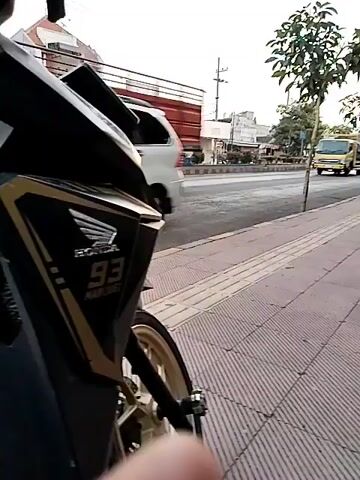 The video shows a close-up of the front of a black and gold motorcycle parked on a sidewalk. The motorcycle has the number 93 and the Honda logo visible. The sidewalk is made of patterned concrete. In the background, there is a street with traffic, including a yellow truck, several cars, and other motorcycles. Buildings line the street, and trees are planted along the sidewalk. The sky is overcast, suggesting it may be morning or late afternoon. The camera is positioned at a low angle, focusing on the motorcycle and the street scene beyond. The video is shot in portrait mode.