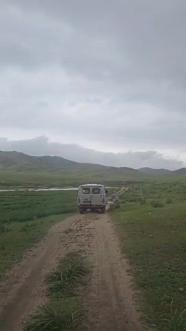 The video shows a white van driving on a dirt road through a grassy field. There are hills in the background and a cloudy sky. The van appears to be moving away from the camera. The road is rutted and muddy in some places. The grass is green and lush. The overall impression is of a remote, undeveloped landscape.