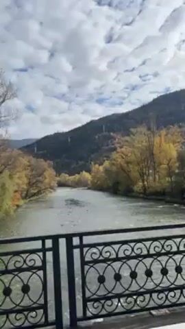 The video shows a view of a river from a bridge. The river is wide and flows between trees with yellow foliage. A mountain covered in green and yellow trees rises in the background under a cloudy sky. The camera moves slightly, capturing different angles of the scene through the ornate iron railing of the bridge. A lamppost is also visible beside the bridge.