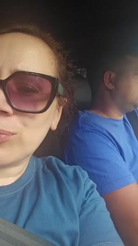 A woman with brown hair wearing large pink-tinted sunglasses and a blue t-shirt is in the front passenger seat of a car. She is looking directly at the camera. Behind her, a man wearing a blue t-shirt is in the driver's seat, looking forward. The interior of the car is visible, with a seatbelt across the woman's chest. The background outside the car is blurred, suggesting movement.
