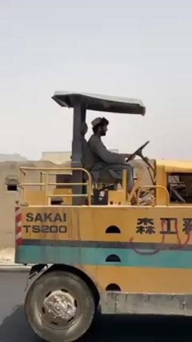A man operates a yellow and blue road roller under a bright, hazy sky. He is seated in an open-air driver's cabin with a small canopy providing shade. He wears a light-colored, long-sleeved garment, possibly a shalwar kameez, and a traditional light-colored cap, possibly a pakol. He has a dark beard and appears focused on driving the machinery.

The road roller is prominently marked with "SAKAI TS200" on its side. Below this text, a horizontal blue-green stripe runs across the machine. Further along the side, Japanese characters (森 工務店, likely "Mori Kōmu-ten" or "Mori Construction Shop") are visible, partially obscured by red spray paint graffiti. The machine is moving slowly forward, suggesting it is engaged in road construction or compacting a surface.

The background shows a dusty, arid environment with several simple, light-colored buildings, possibly made of concrete block or mud-brick, under a wide expanse of pale sky. Distant mountains or hills are visible through the atmospheric haze. The strong overhead sun indicates it is midday, and the general appearance of the landscape suggests a dry, hot climate. The distinct sound of the road roller's engine is clearly audible throughout the video. The scene depicts a typical day of construction work in what appears to be a developing region, consistent with the provided location context of Kandahar, Afghanistan.