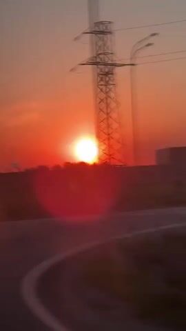 The video shows a vivid red sunset behind a treeline, observed from a moving vehicle.  Power lines stretch across the upper portion of the frame, and a silhouette of a factory emitting smoke is visible on the horizon. A road appears in the foreground. A car briefly passes by the camera.