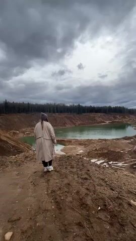 A woman in a tan trench coat and a dark beanie stands on a muddy bank overlooking a green lake. She is turned away from the camera at first, then turns her head to smile briefly. She raises her arms above her head before lowering them. The sky is overcast and cloudy. A line of trees is visible in the background. The ground is dirt and mud.