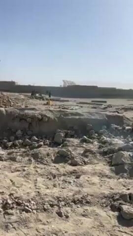 The video shows a desolate, dusty landscape with remnants of destroyed buildings. The foreground is a rough, uneven terrain covered with rocks and dirt. In the background, a partially demolished building stands, showing its bare structure and broken walls. There's a group of men, dressed in traditional clothing, walking toward the building. The sky is clear and blue, suggesting a sunny day. A lone tree stands to the side, adding a touch of life to the otherwise barren scene.