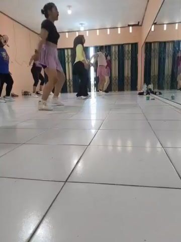 A group of women are in a dance studio, practicing a routine. The studio has a tiled floor and mirrors along one wall. The women are dressed in athletic wear, including skirts and leggings. They are all facing the mirrors and appear to be following an instructor. The lighting in the room is bright, and there are some decorative curtains on the wall behind the women.