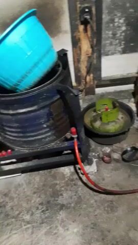 The video shows an outdoor area with a concrete floor and a rough, textured wall in the background. In the foreground, there is a blue barrel that appears to be used as a stove or heating apparatus. A gas cylinder with a red regulator and hose is connected to the barrel. Next to the barrel is another large drum, possibly for water or storage, with a teal-colored lid. To the right of the blue barrel, there is a large wok or cooking pot placed over a fire pit, which appears to be made of bricks and metal. The area around the cooking setup shows signs of use, with ash and debris scattered on the ground. The lighting is dim, suggesting it might be evening or an indoor/covered space. There are no people visible in the video.