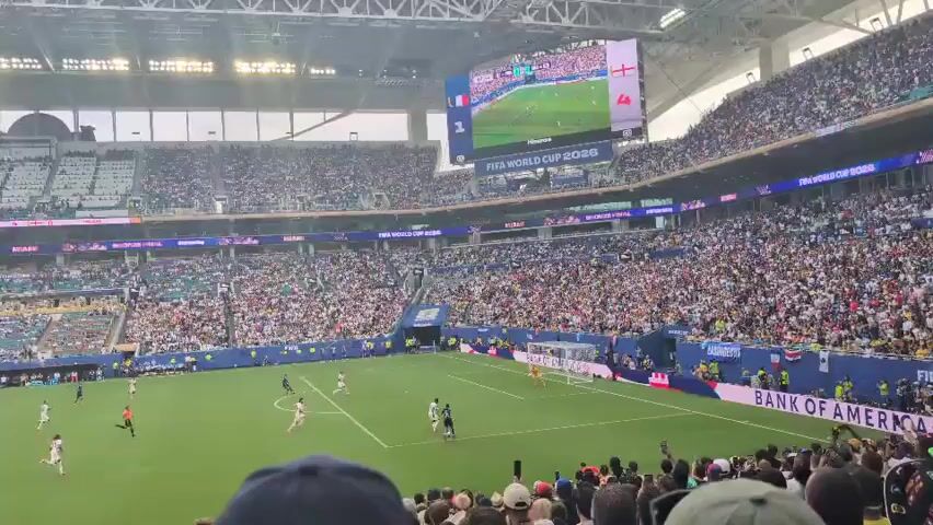 A wide-angle shot shows a soccer stadium filled with a large crowd. On the green field, several players in white and red uniforms are engaged in a soccer match. A large screen in the background displays the FIFA World Cup 2026 logo. The spectators are seated in tiered stands that surround the field. The atmosphere appears lively, with many people holding up phones, presumably to record the event. The stadium is well-lit, indicating it's daytime or under bright artificial lighting. The crowd is diverse, with people of various ages and backgrounds. The perspective is from a spectator in the stands, looking down at the field.
