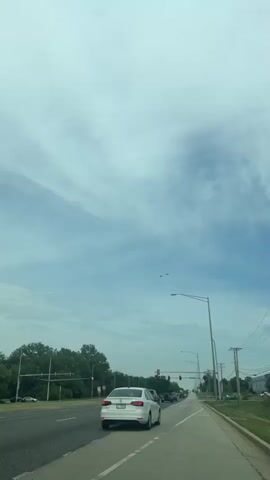 The video shows two small planes flying in formation in a slightly cloudy sky. The video is taken from the perspective of someone inside a car that is stopped at a traffic light on a road lined with trees and streetlights. Other cars are visible on the road. There is some speech in the background.