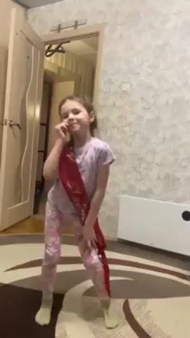 A young girl, perhaps around 6-8 years old, stands in a room and poses for the camera. She is wearing a light-colored pajama set with a pattern of pink and white shapes, and white socks. A red sash with gold lettering is draped across her body, the words appearing to read "Выпускник" (likely Russian for "Graduate"). The room has patterned wallpaper in shades of gray and white. There is a beige door on the left and an open doorway behind her. A white radiator is mounted on the wall to her right. The floor is covered with a tan rug with dark brown concentric oval shapes. The lighting in the room appears to be soft and diffuse.