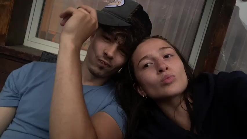 A man and a woman are in the foreground, with the man on the left and the woman on the right. They appear to be in an indoor setting, possibly a room with a window and curtains visible in the background. The man is wearing a blue t-shirt and a dark baseball cap. He is looking at the camera with a slight frown and his left arm is raised, with his hand resting on the brim of his cap. The woman is wearing a dark hooded sweatshirt. She is looking directly at the camera with her tongue sticking out slightly. Her right arm is visible, with her hand resting on her chest. The lighting is dim.
